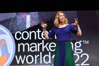 White woman with blonde hair wearing a navy blue shirt and green and navy pleated skirt on stage.